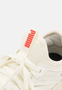 Białe siatkowe sneakersy z teksturowanymi bokami, płaskimi sznurówkami i wyraźną czerwoną metką "Puma". Posiadają wyściełany kołnierz i minimalistyczny design.