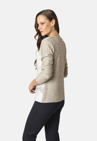 Una mujer con cabello largo castaño lleva un suéter de patrones beige y blanco y pantalones negros, de pie en una pose de tres cuartos contra un fondo blanco.