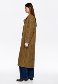 Abrigo de espiga marrón con mangas largas, corte oversize y cuello pico, combinado con jeans oscuros de pierna ancha y botas de tacón negras.
