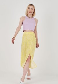 Lila ribbad crop top och en gul blommönstrad wrapkjol med en sidofissyr, kombinerad med vita sandaler och guldchain-accessoarer.