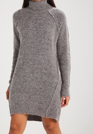 Kobieta ubrana w długorękawową szarą dzianinową sukienkę z golfem o lekko asymetrycznym dole na jednolitym tle.