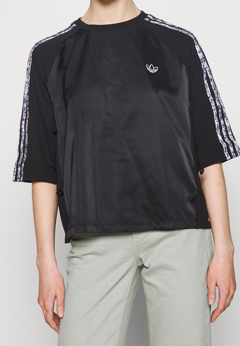 Persona con camiseta negra de manga corta Adidas con tres rayas blancas estampadas en las mangas y pantalones de color claro.