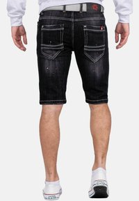 Corti di jeans neri con una finitura consumata, cuciture visibili e cinque tasche. Indossati con sneaker bianche, che mostrano i peli delle gambe e il tono della pelle.