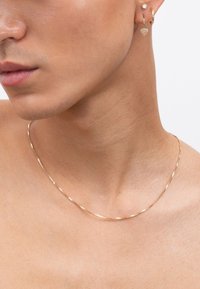 Goldene Kette mit strukturiertem Oberflächenfinish, kombiniert mit einem kleinen herzförmigen Ohrring und einem schlichten Creolen-Ohrring, vor Haut präsentiert.