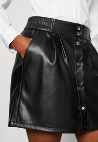 Jupe en cuir noir avec une taille élastique froncée, des poches latérales et des clous pyramidaux argentés décoratifs le long de la patte de boutonnage. Texture lisse.