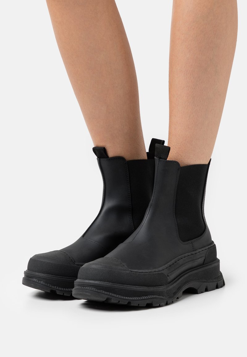 Botas de ankle pretas feitas de couro liso, com painéis laterais elásticos, sola grossa e um padrão resistente para tração.