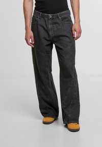 Black denim wide-leg pants with curved seam details, five pockets, and a button fly; worn with yellow shoes.
