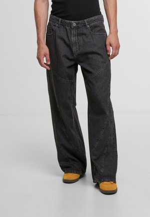 Schwarze Denim-Hose im weiten Schnitt mit gekrümmten Nahtdetails, fünf Taschen und Knopfverschluss; getragen mit gelben Schuhen.