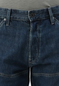 Σκούρο μπλε τζιν παντελόνι με ορατές ραφές, κλείσιμο με κουμπί, θηλύκια ζώνης και μεταλλικά τρουκς στις μπροστινές τσέπες, φορεμένο με γκρι μπλουζάκι.