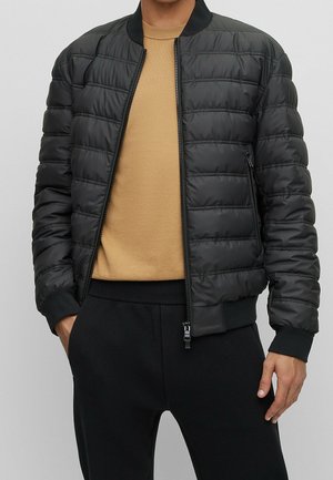 Homme portant une veste bomber noire matelassée par-dessus un pull col rond beige et un pantalon noir, debout avec les mains près des poches.