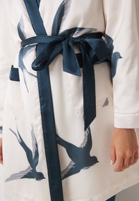 Robe di colore chiaro con una cintura in raso blu scuro. Presenta un motivo di uccelli in volo in grigio e blu navy. Include due tasche frontali.