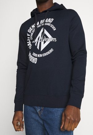 Mann trägt einen marineblauen Hoodie mit weißem, kreisförmigem „Quality Denim Brand“-Schriftzug und Grafik, kombiniert mit blauen Jeans, Hände entspannt an den Seiten.