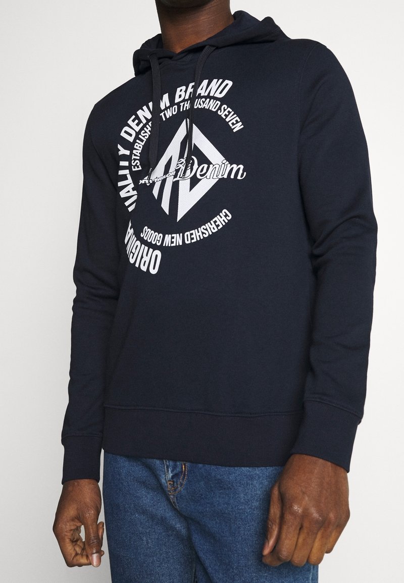 Homme portant un sweat à capuche bleu marine avec un texte circulaire blanc « Quality Denim Brand » et un graphisme, assorti à un jean bleu, mains détendues sur les côtés.