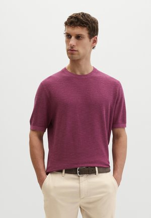 Junger Mann trägt einen kurzärmligen, violetten Strickpullover, der in beige Hosen mit braunem Gürtel gesteckt ist, steht mit den Händen in den Taschen.