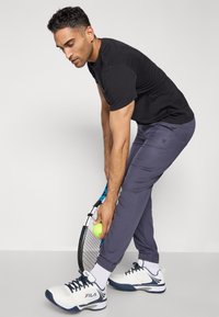 Černé tričko s krátkým rukávem, šedé sportovní kalhoty s elastickými manžetami a bílé tenisky. Drží tenisovou raketu a žlutý tenisový míček.