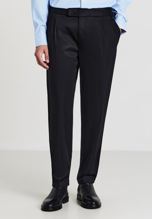 Pantalons noirs ajustés avec une texture lisse, présentant une taille plate, des poches latérales et une passante de ceinture. Portés avec des chaussures de ville noires.