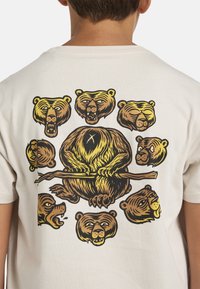 Kerman lyhythihainen T-paita, jossa on yksityiskohtainen karhugrafiikka ruskeassa ja keltaisessa värissä. T-paidassa on useita karhun päitä keskellä istuvan karhun ympärillä.