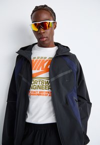 Black sports jacket with navy accents, featuring a zippered pocket and reflective details. Underneath, a white t-shirt with orange and green text.