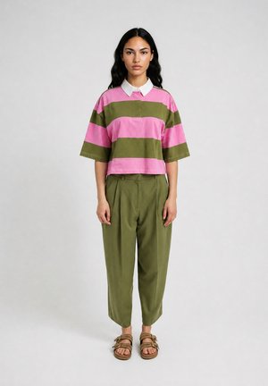Woman with long dark hair wears a pink and green striped shirt with white collar, olive green pants, and tan sandals, standing against plain background.