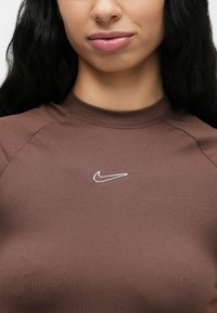 Καφέ εφαρμοστό αθλητικό τοπ με ψηλό λαιμόκοψη. Διαθέτει μικρό ασημί λογότυπο Nike στο στήθος. Ομαλό, ελαστικό ύφασμα με αδιάκοπο σχέδιο.