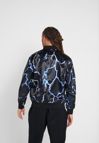 Musta satiininen bomber-takki, jossa on rohkea sininen salaman kuvio, resorikahleet ja -helma sekä klassinen kaulussuunnittelu.