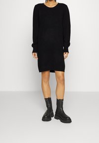Robe pull noire en maille avec manches longues, col rond et poignets côtelés ; associée à des bottes noires à semelles épaisses.