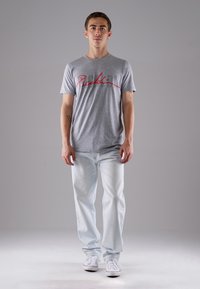 Graues Baumwoll-T-Shirt mit rotem, geschwungenem Text "Piccolo", kombiniert mit hellblauen Jeans und weißen Sneakers. Modell steht vor einem grauen Hintergrund.