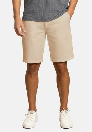 Bermudas color caqui hechas de una mezcla de algodón suave, que presentan un corte holgado, una parte frontal plana y bolsillos laterales, combinadas con zapatillas blancas de cordones.