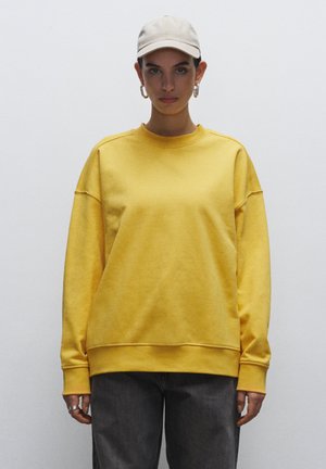 Yellow cotton sweatshirt with long sleeves, crew neck, and dropped shoulders. Paired with gray denim pants and a beige cap.