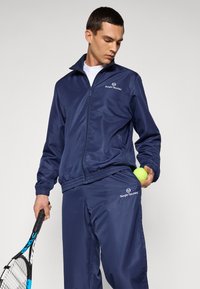Marineblaues Trainingsanzug aus glattem, leichtem Material; umfasst eine Zip-Jacke mit Logo und Jogginghosen mit Kordelzug, die einen Tennisball hält.