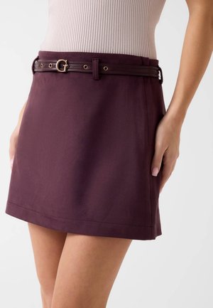 Femme portant une jupe violet foncé avec une fine ceinture marron foncé et un haut côtelé rose clair sur un fond uni.