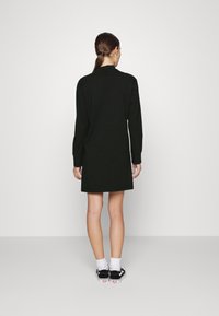 Abito lungo nero a maniche lunghe con collo alto, realizzato in tessuto morbido. Presenta una linea dritta e un design minimalista, abbinato a sneakers nere.