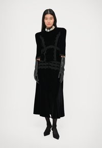 Μαύρο βελούδινο φόρεμα με λεπτομέρειες δαντέλας, μακριά μανίκια και εφαρμοστό κορσέ. Συνδυάζεται με μαύρα γάντια και τακούνια με μυτερή μύτη.