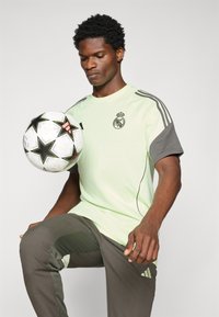 T-shirt de sport vert clair avec des manches gris foncé, arborant un logo. Le modèle tient un ballon de football noir et blanc décoré d'étoiles.