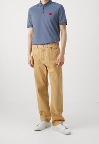 Mörkblå herrpolo med en röd applikation, tillsammans med ljusbeiga byxor och vita sneakers. Slät textur; klassisk passform.