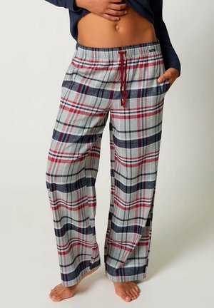 Person wearing loose gray, red, and black plaid pajama pants with a red drawstring, standing barefoot with one hand in the pocket.