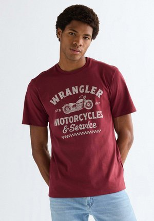 Junger Mann trägt ein weinrotes Wrangler-T-Shirt mit einem Vintage-Motorrad-Design und Text, kombiniert mit hellblauen Jeans.