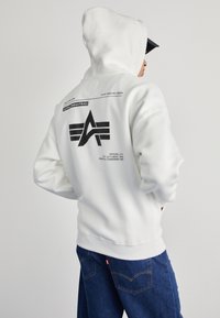 Personne portant un sweat à capuche blanc Alpha Industries avec un logo noir et un texte au dos, associé à un jean bleu et une casquette noire.