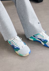Tenisi albi cu accente albastre și verzi, superior din plasă și șireturi albe. Pantaloni de trening gri cu un logo pe tiv. Fundal neutru.