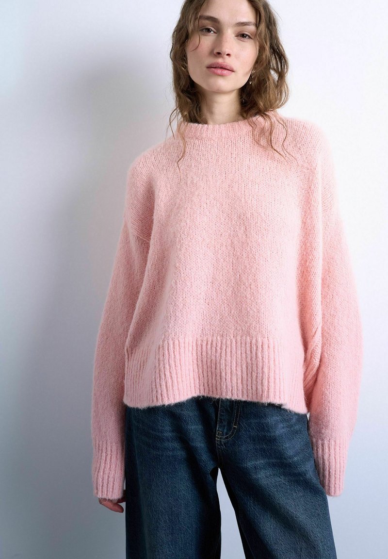 Femme portant un pull ample en tricot rose pâle doux et un jean bleu foncé, debout contre un fond gris clair.