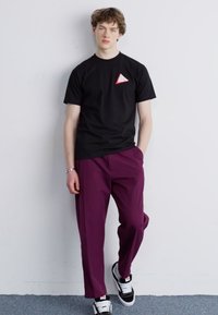 Camiseta negra con un pequeño logo de triángulo rojo, combinada con pantalones morados de corte holgado. Zapatillas negras y blancas completan el look.