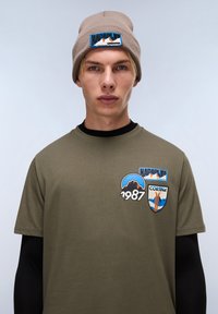 Olivově zelené tričko s černými dlouhými rukávy, s barevnými vyšívanými nášivkami včetně hor a textů "Napapijri" a "Cortina."