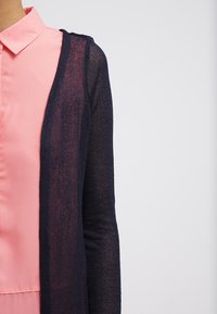 Marinblå, stickad kofta med långa ärmar, halvgenomskinlig textur. Kombinerad med en ljusrosa skjorta med krage underneath.