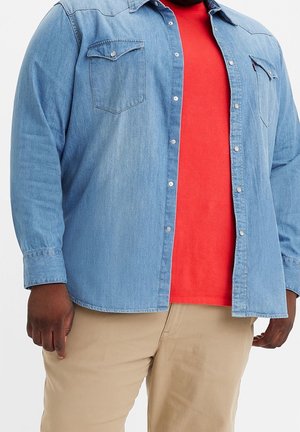 Mann trägt ein rotes T-Shirt, ein hellblaues Jeans-Hemd mit Knöpfen und beige Hosen, steht vor einem schlichten weißen Hintergrund.