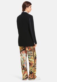 Black blazer with contrasting side panels, paired with colorful, patterned wide-leg trousers featuring tropical and animal motifs. White footwear.