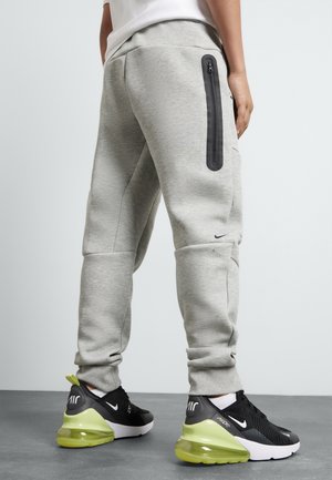 Personne portant un pantalon de jogging gris clair avec une poche latérale zippée noire et des baskets Nike Air noires avec des semelles jaunes.