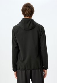 Chaqueta negra con capucha de textura suave, puños elásticos y diseño de ventilación en la parte posterior. Presenta un dobladillo recto y un estilo minimalista.