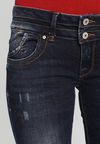 Jeans de mezclilla azul oscuro con cintura alta, que presentan dos botones en la parte delantera, un ligero desgaste y detalles de costura en color naranja.