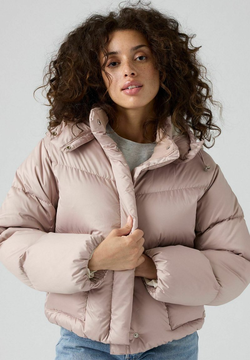 Chaqueta acolchada de color rosa claro con un alto cuello, grandes mangas voluminosas y cierre de botones. Textura suave con un bolsillo delantero en el lado izquierdo.