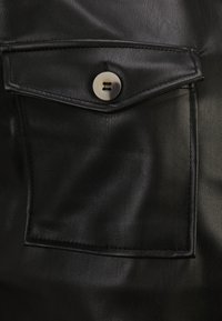 Mustapahkainen nahkalaukku napin suljennalla, jossa on sileä pinta ja ommellut reunat, sijoitettuna suuremmalle nahkapinnalle.